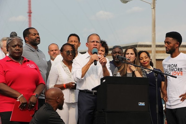 Anthony Brown speaks at a podium during an outdoor event, surrounded by a group of supporters.
