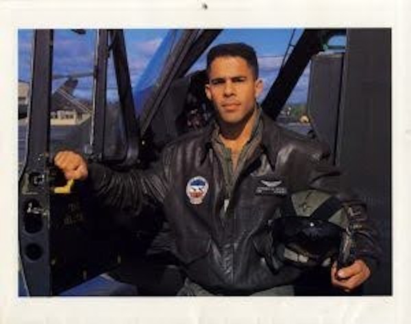 Younger Anthony Brown standing in front of a military aircraft wearing a flight jacket and holding a helmet