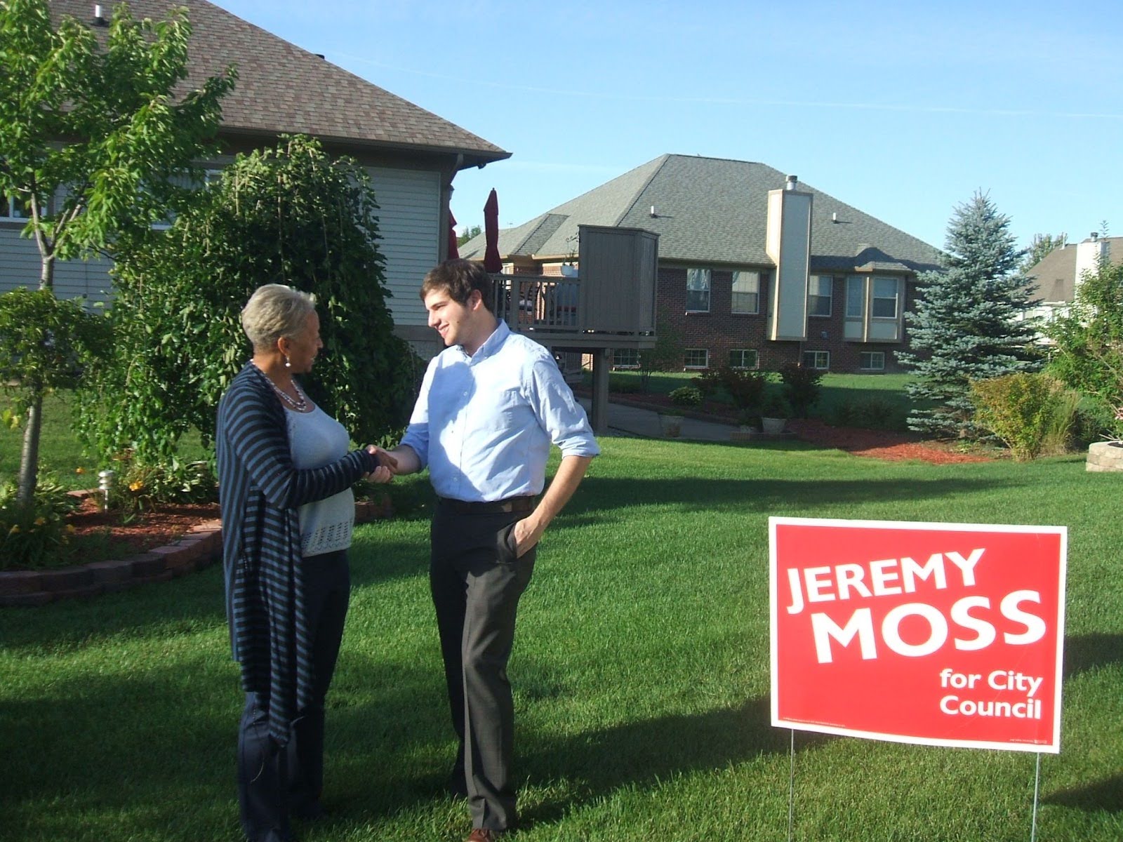 Younger Jeremy Moss campaigning for city council, shaking hands with a resident beside a campaign sign.